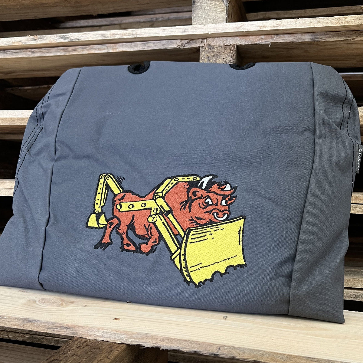 A dark gray canvas folded seat cover featuring an embroidered graphic of a red wild boar wearing a yellow construction helmet and holding a yellow shovel, displayed on a rustic woden pallet.