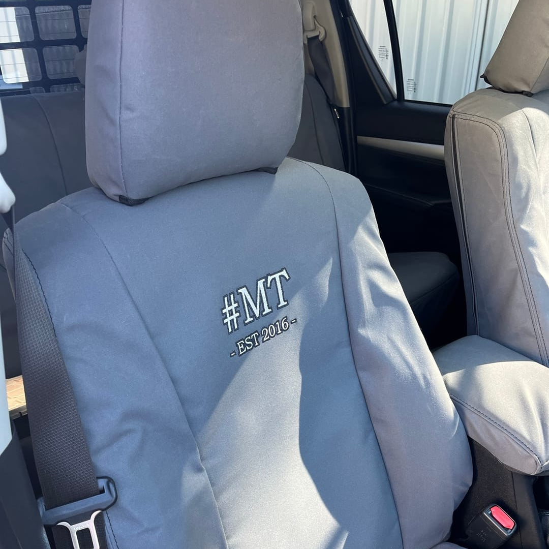 A gray car seat cover installed on a vehicle's front passenger seat, featuring the embroidered text '#MT EST 2016' on the backrest.